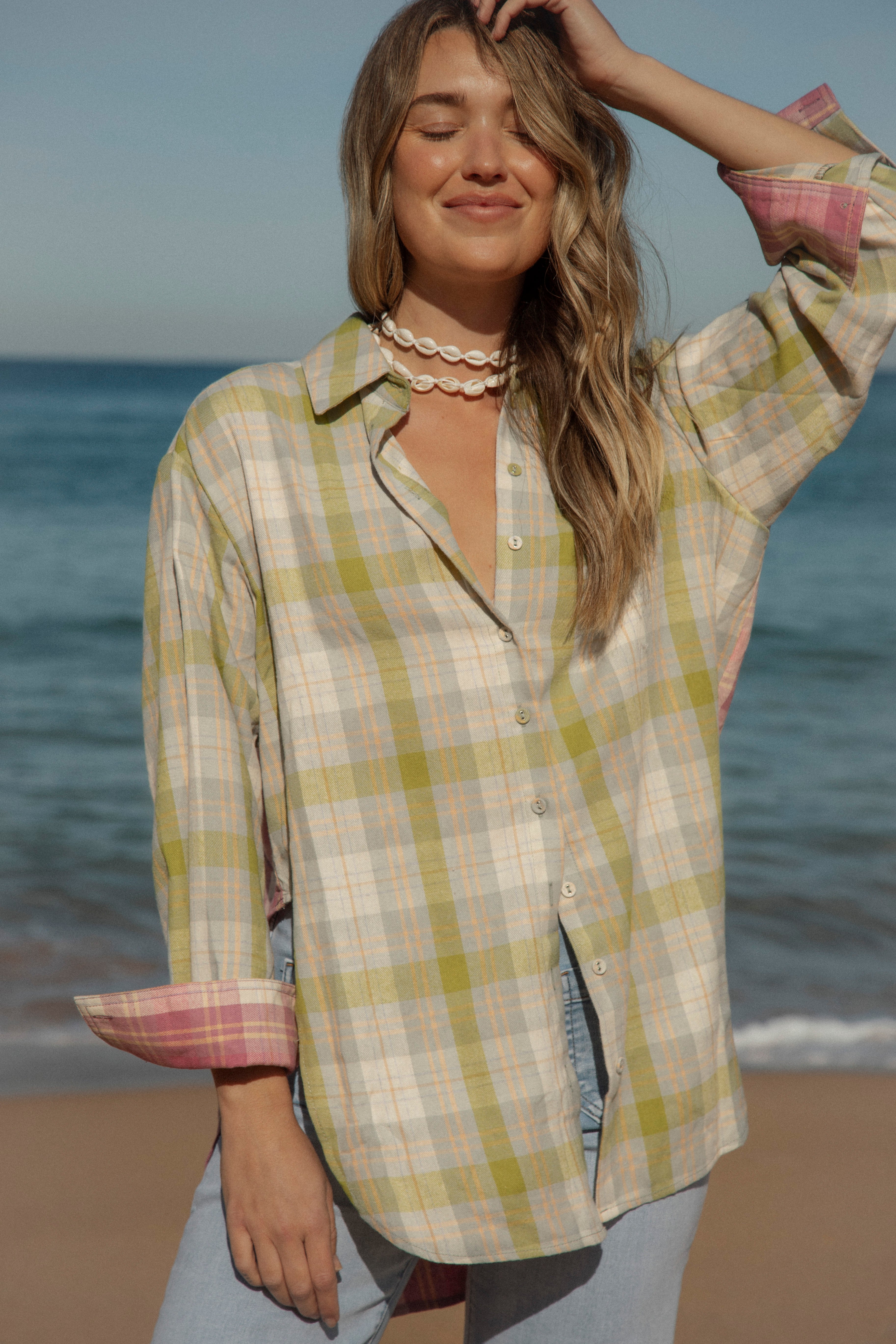 Oversized plaid button-up shirt in olive, peach, and beige with contrast pink cuffs, styled with shell necklace at the beach