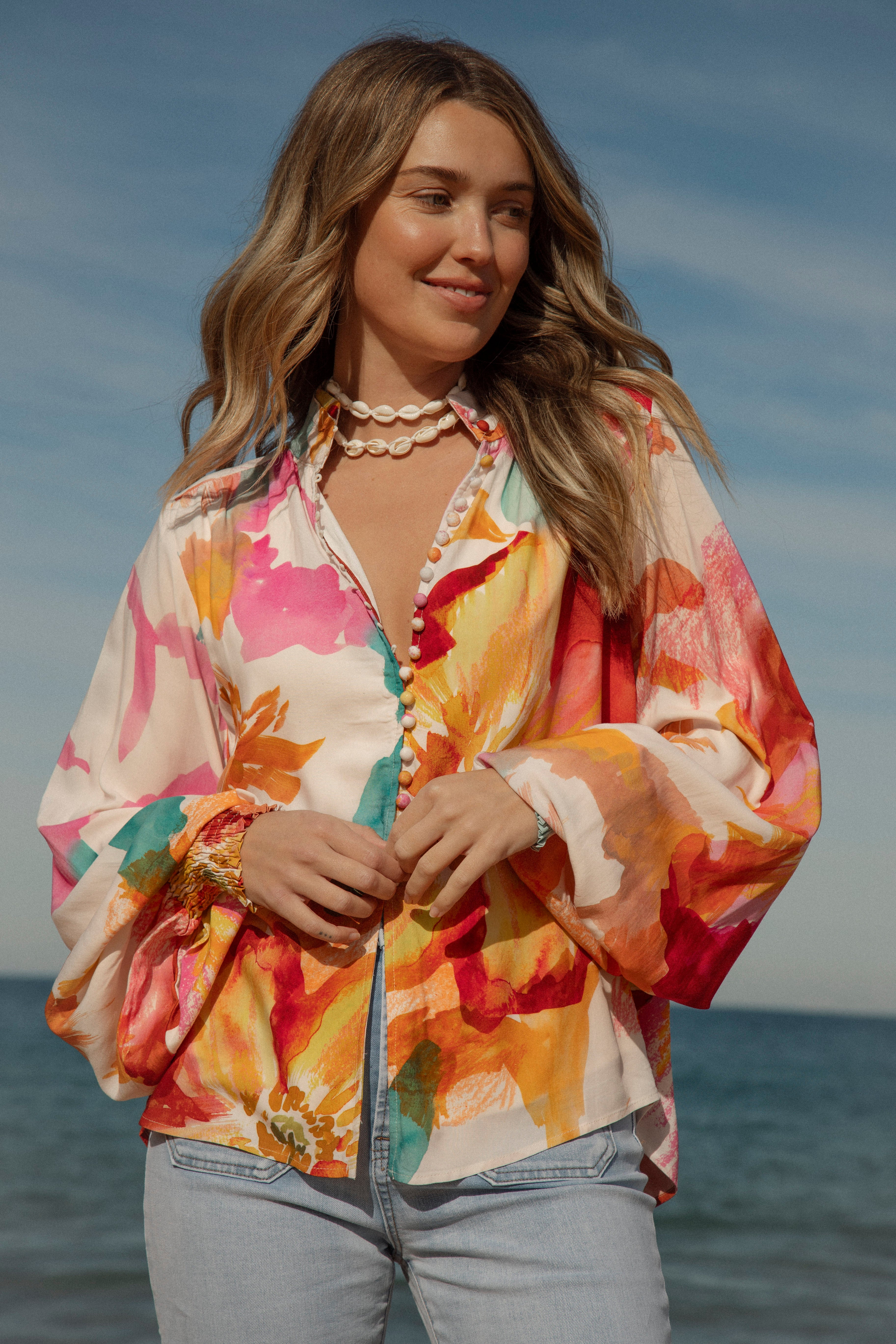 Woman wearing a vibrant floral blouse with puffed long sleeves and colorful buttons, standing against a blue sky and ocean background. The lightweight, flowy top features bold orange, pink, and yellow watercolor-style floral prints. Styled with light-wash high-rise jeans and a shell necklace for a beachy bohemian look.