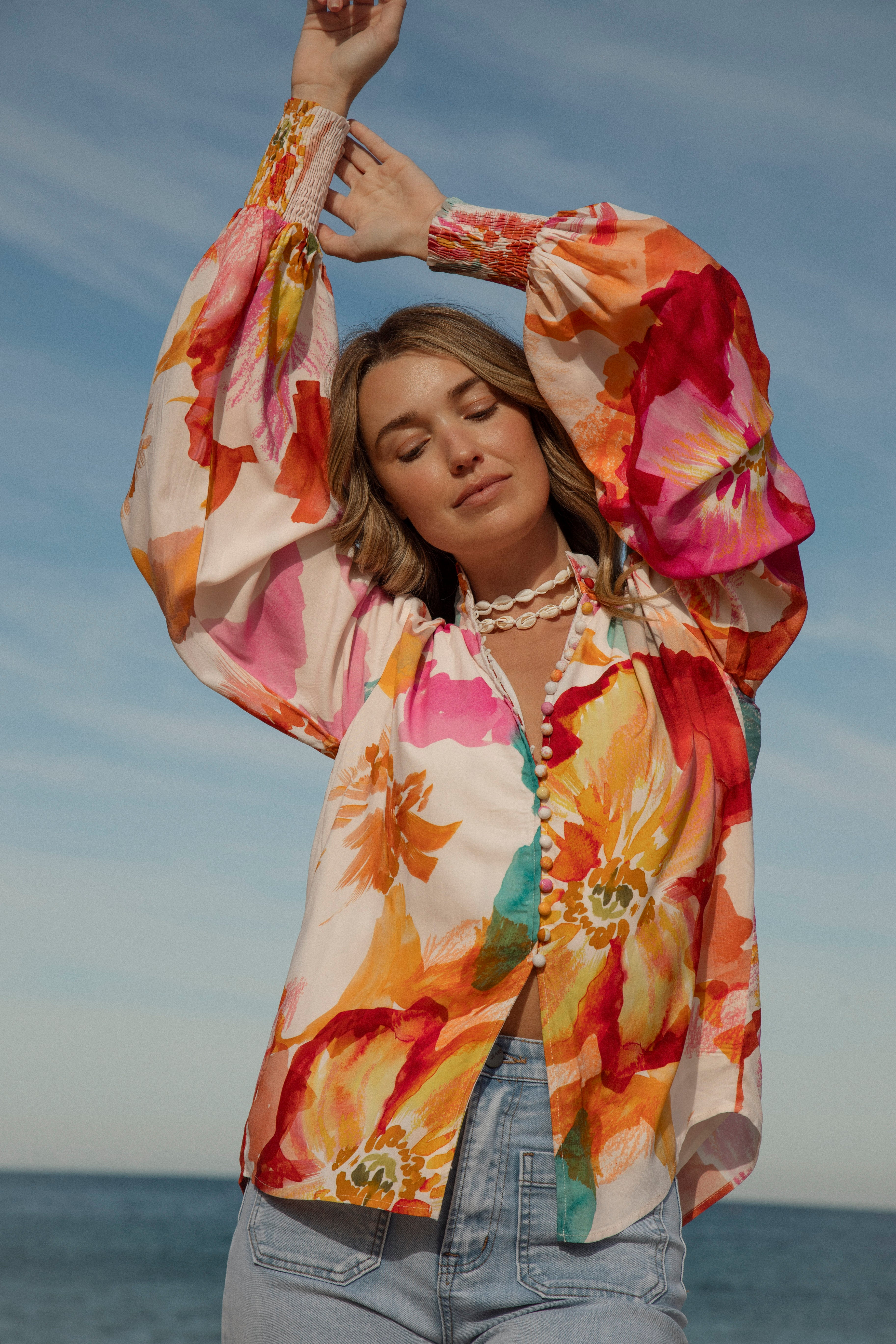 Woman wearing a vibrant floral blouse with puffed long sleeves and colorful buttons, standing against a blue sky and ocean background. The lightweight, flowy top features bold orange, pink, and yellow watercolor-style floral prints. Styled with light-wash high-rise jeans and a shell necklace for a beachy bohemian look.