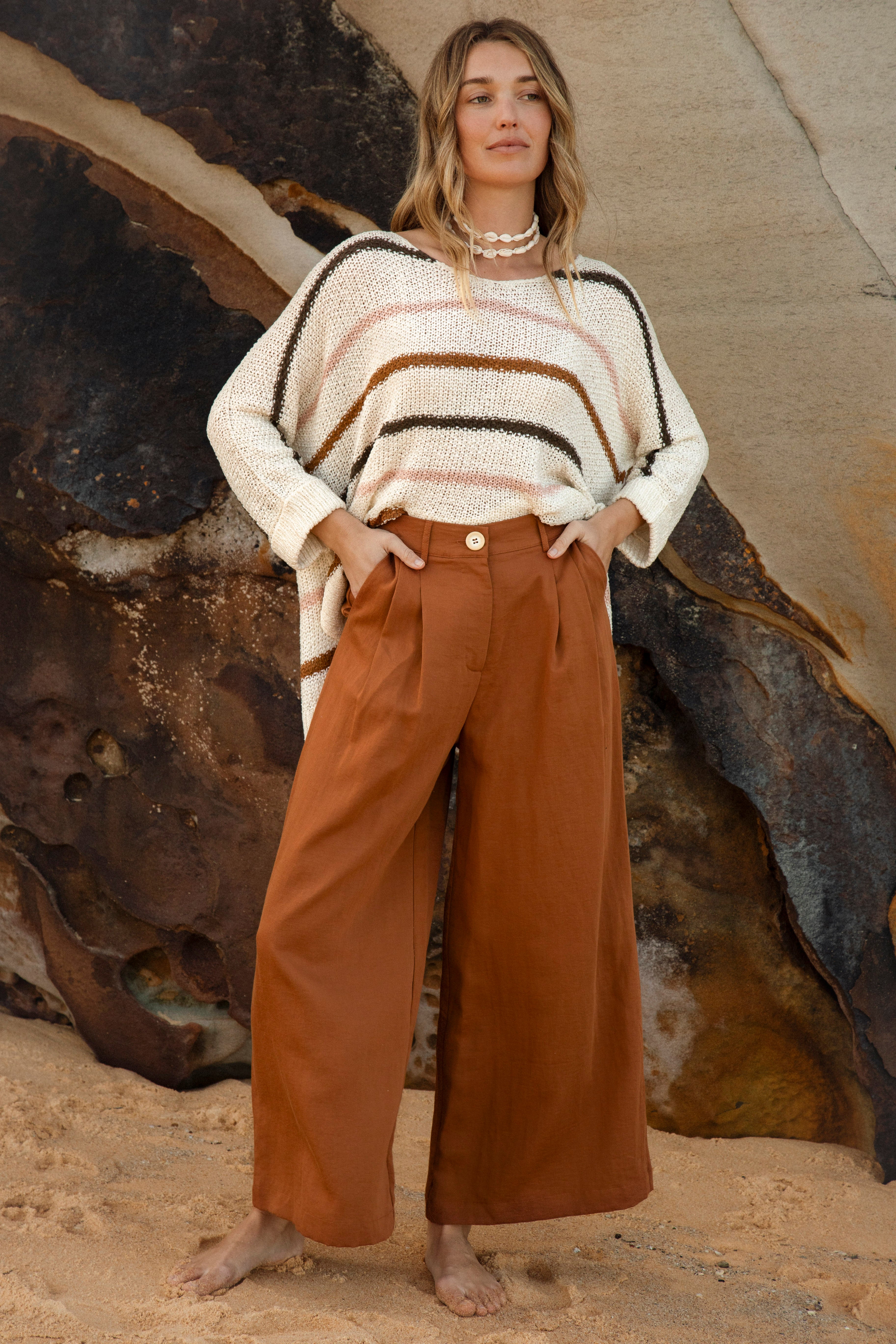 Woman wearing high-waisted terracotta wide-leg trousers with pleated front and side pockets, styled casually on the beach with striped knit top