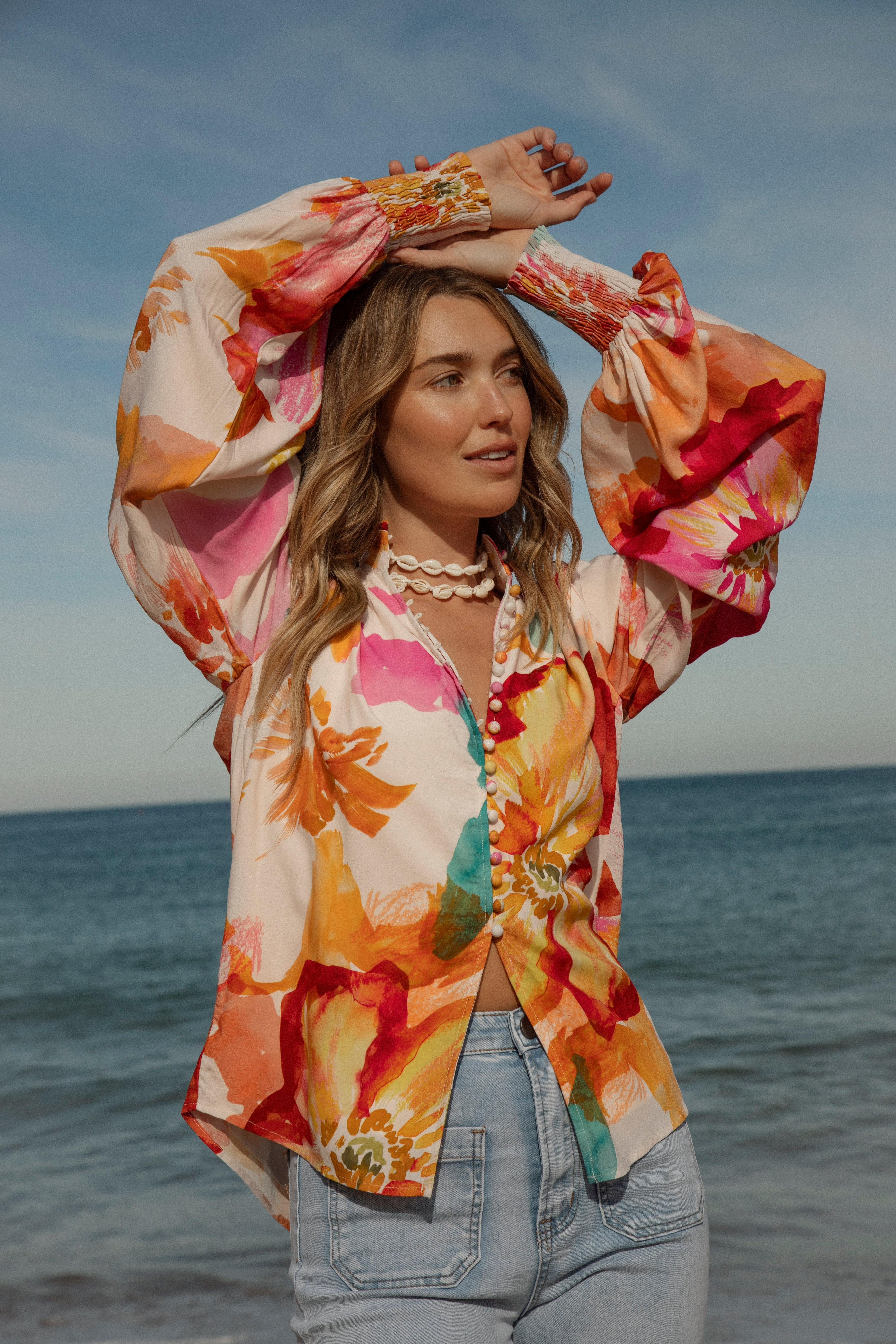 Woman wearing a vibrant floral blouse with puffed long sleeves and colorful buttons, standing against a blue sky and ocean background. The lightweight, flowy top features bold orange, pink, and yellow watercolor-style floral prints. Styled with light-wash high-rise jeans and a shell necklace for a beachy bohemian look.