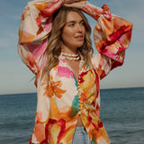 Woman wearing a vibrant floral blouse with puffed long sleeves and colorful buttons, standing against a blue sky and ocean background. The lightweight, flowy top features bold orange, pink, and yellow watercolor-style floral prints. Styled with light-wash high-rise jeans and a shell necklace for a beachy bohemian look.