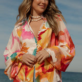Woman wearing a vibrant floral blouse with puffed long sleeves and colorful buttons, standing against a blue sky and ocean background. The lightweight, flowy top features bold orange, pink, and yellow watercolor-style floral prints. Styled with light-wash high-rise jeans and a shell necklace for a beachy bohemian look.