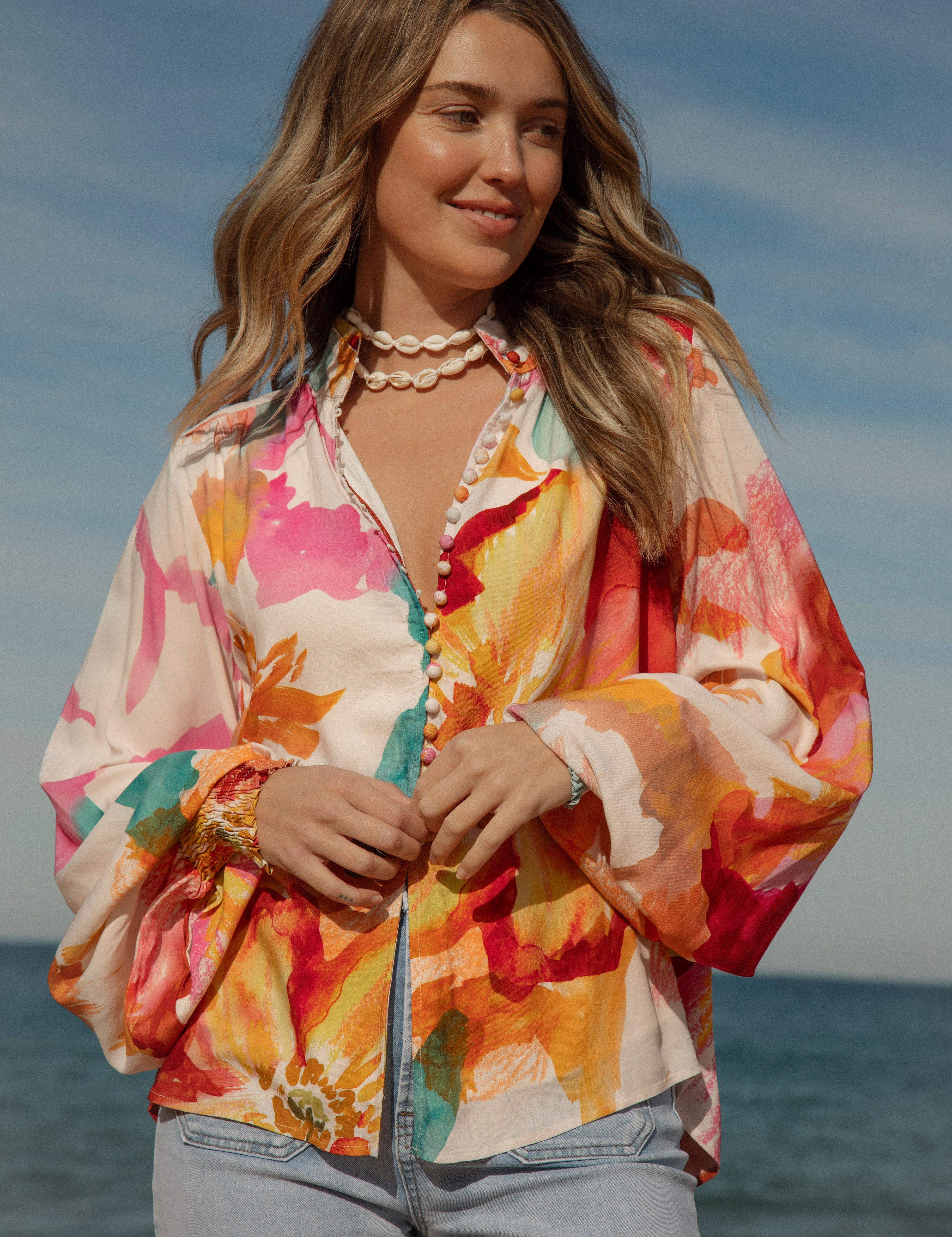 Woman wearing a vibrant floral blouse with puffed long sleeves and colorful buttons, standing against a blue sky and ocean background. The lightweight, flowy top features bold orange, pink, and yellow watercolor-style floral prints. Styled with light-wash high-rise jeans and a shell necklace for a beachy bohemian look.