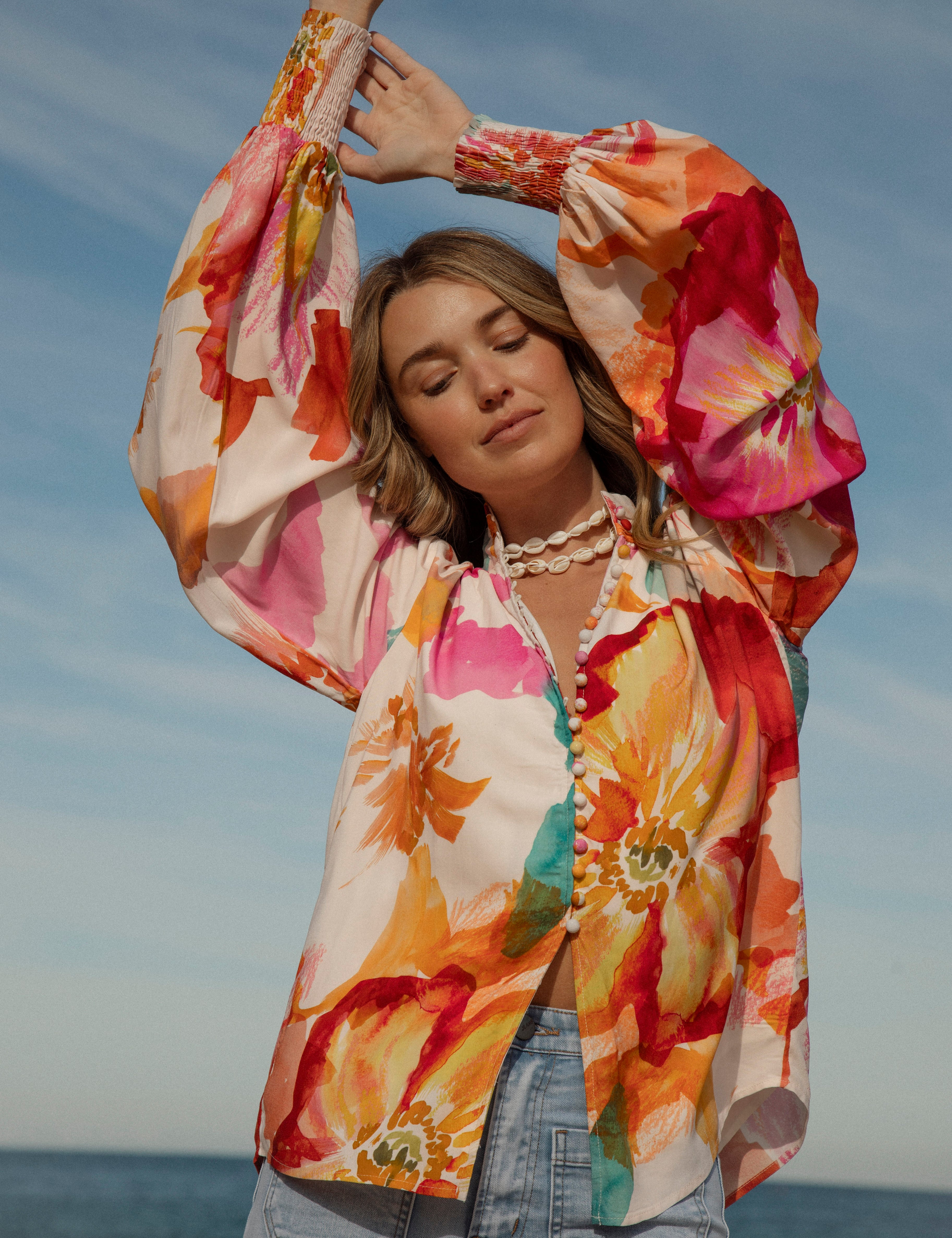 Woman wearing a vibrant floral blouse with puffed long sleeves and colorful buttons, standing against a blue sky and ocean background. The lightweight, flowy top features bold orange, pink, and yellow watercolor-style floral prints. Styled with light-wash high-rise jeans and a shell necklace for a beachy bohemian look.