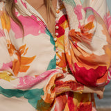 Woman wearing a vibrant floral blouse with puffed long sleeves and colorful buttons, standing against a blue sky and ocean background. The lightweight, flowy top features bold orange, pink, and yellow watercolor-style floral prints. Styled with light-wash high-rise jeans and a shell necklace for a beachy bohemian look.
