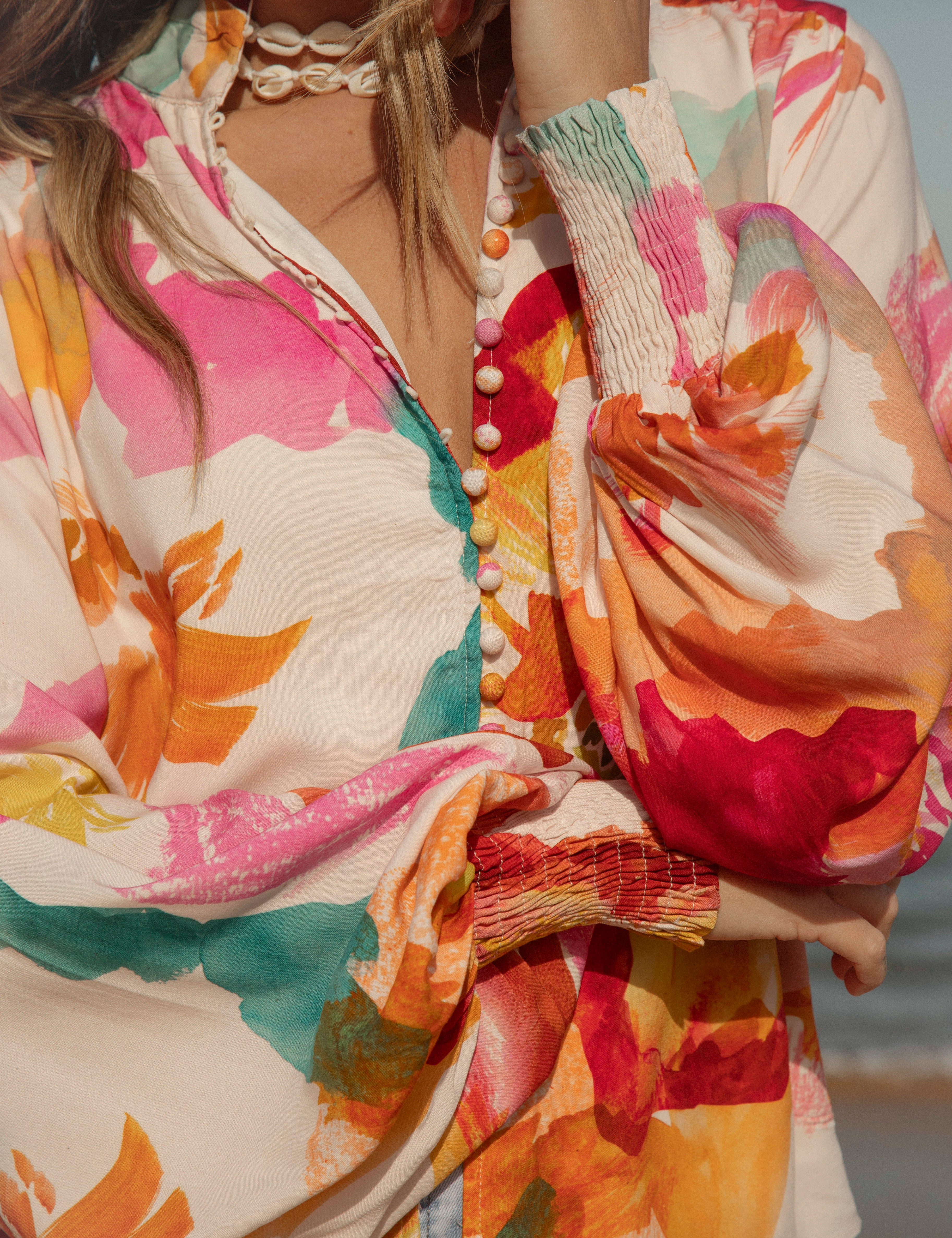 Woman wearing a vibrant floral blouse with puffed long sleeves and colorful buttons, standing against a blue sky and ocean background. The lightweight, flowy top features bold orange, pink, and yellow watercolor-style floral prints. Styled with light-wash high-rise jeans and a shell necklace for a beachy bohemian look.