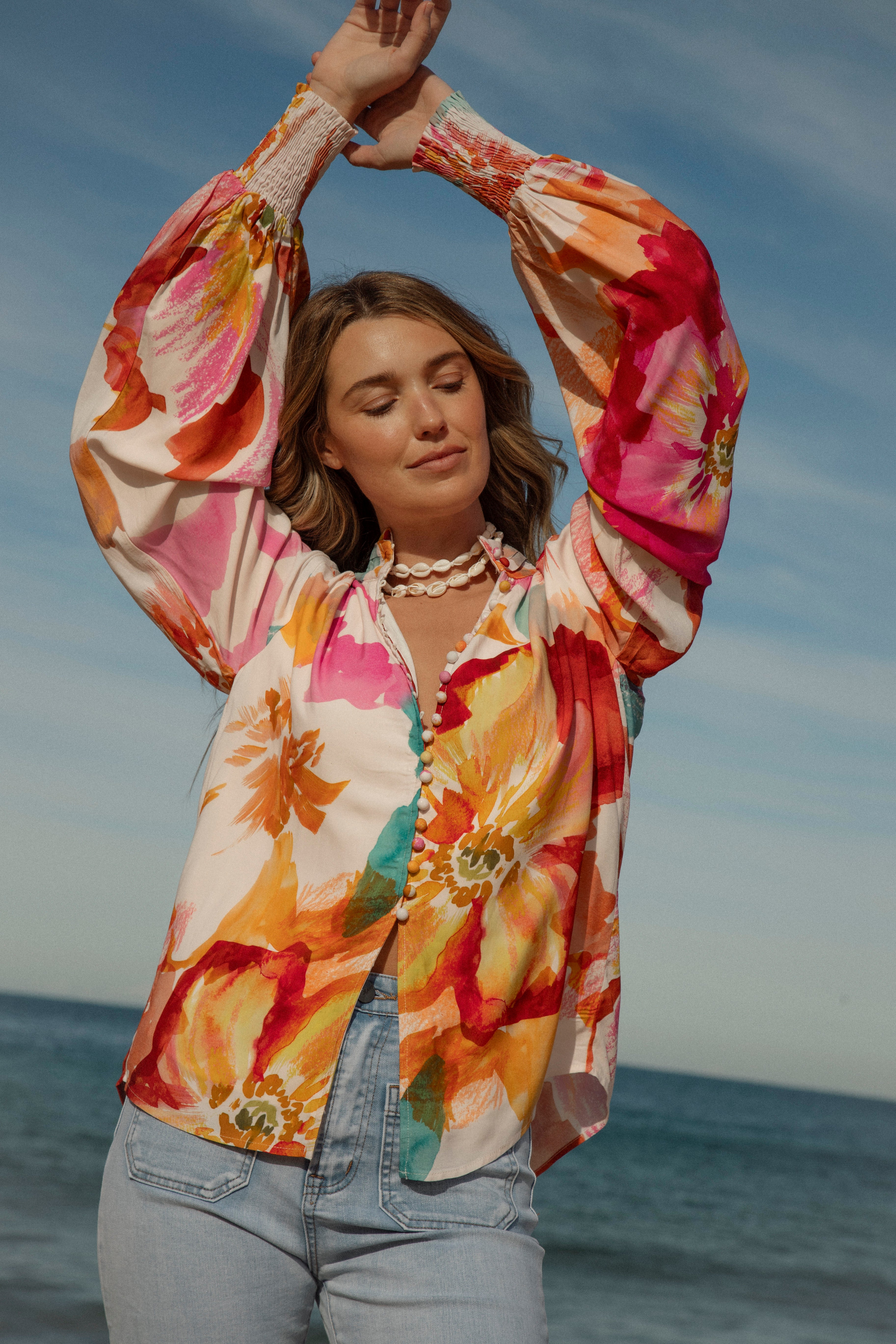 Woman wearing a vibrant floral blouse with puffed long sleeves and colorful buttons, standing against a blue sky and ocean background. The lightweight, flowy top features bold orange, pink, and yellow watercolor-style floral prints. Styled with light-wash high-rise jeans and a shell necklace for a beachy bohemian look.