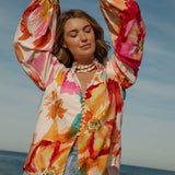 Woman wearing a vibrant floral blouse with puffed long sleeves and colorful buttons, standing against a blue sky and ocean background. The lightweight, flowy top features bold orange, pink, and yellow watercolor-style floral prints. Styled with light-wash high-rise jeans and a shell necklace for a beachy bohemian look.