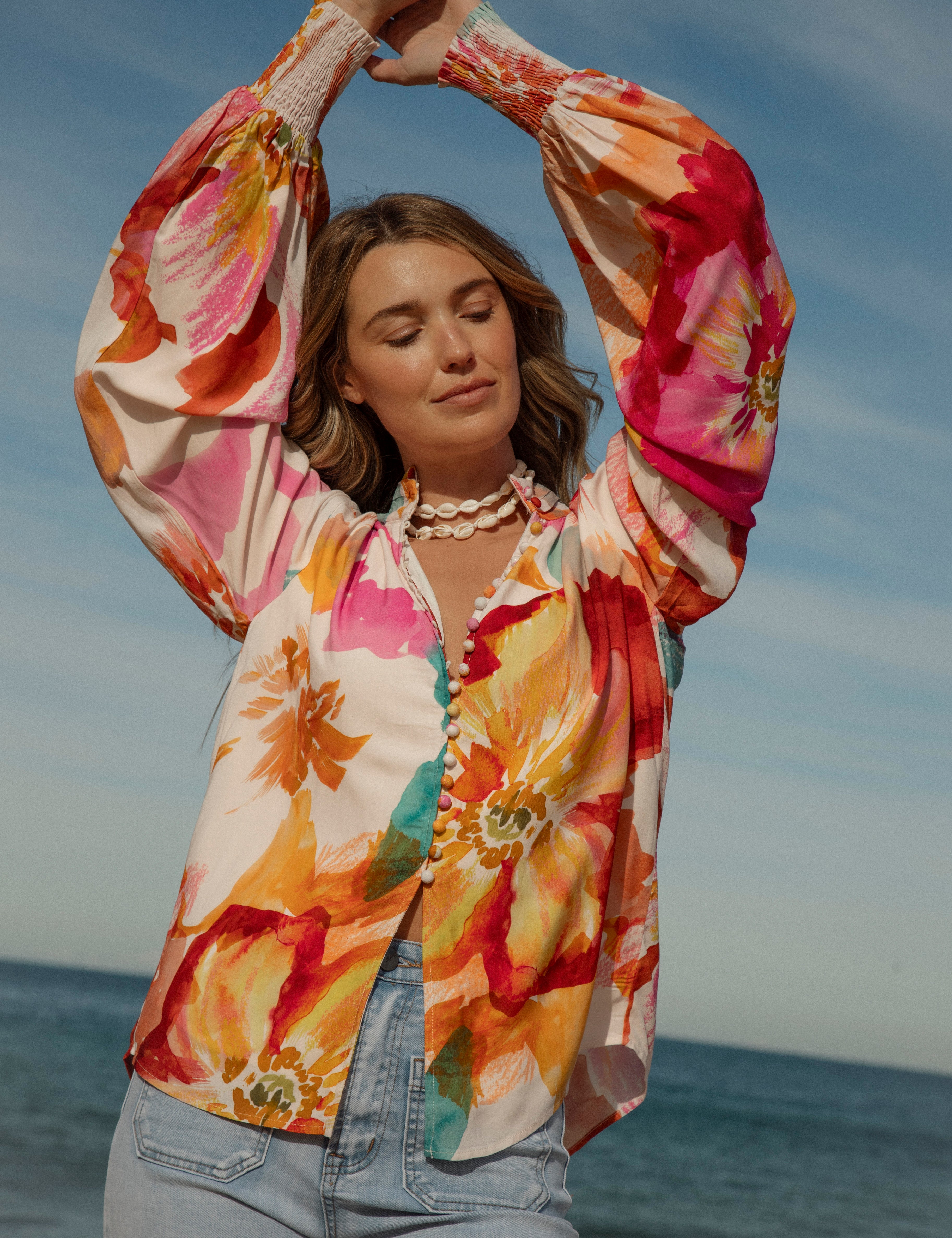 Woman wearing a vibrant floral blouse with puffed long sleeves and colorful buttons, standing against a blue sky and ocean background. The lightweight, flowy top features bold orange, pink, and yellow watercolor-style floral prints. Styled with light-wash high-rise jeans and a shell necklace for a beachy bohemian look.