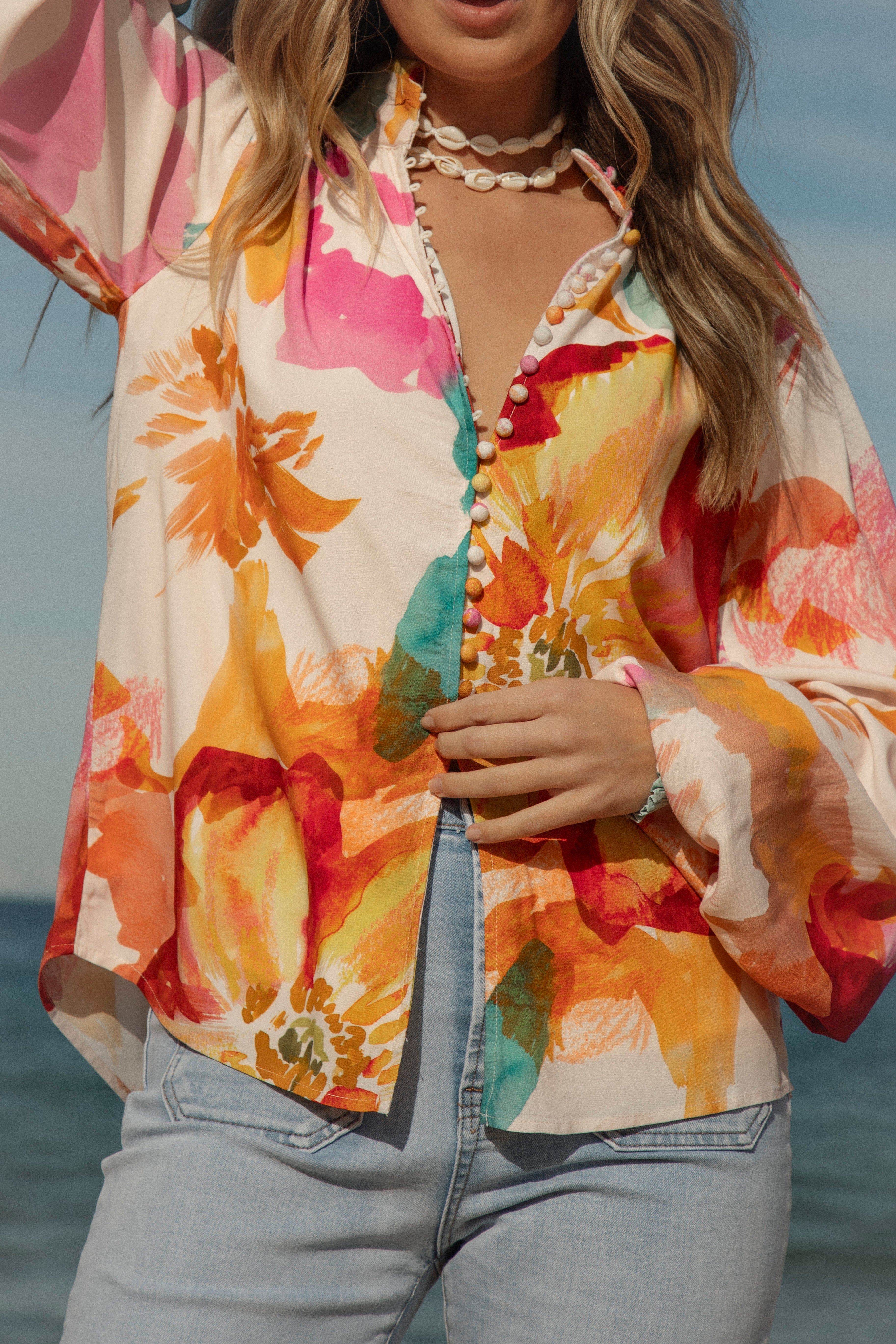 Woman wearing a vibrant floral blouse with puffed long sleeves and colorful buttons, standing against a blue sky and ocean background. The lightweight, flowy top features bold orange, pink, and yellow watercolor-style floral prints. Styled with light-wash high-rise jeans and a shell necklace for a beachy bohemian look.