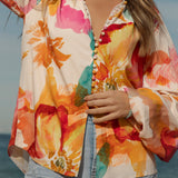 Woman wearing a vibrant floral blouse with puffed long sleeves and colorful buttons, standing against a blue sky and ocean background. The lightweight, flowy top features bold orange, pink, and yellow watercolor-style floral prints. Styled with light-wash high-rise jeans and a shell necklace for a beachy bohemian look.