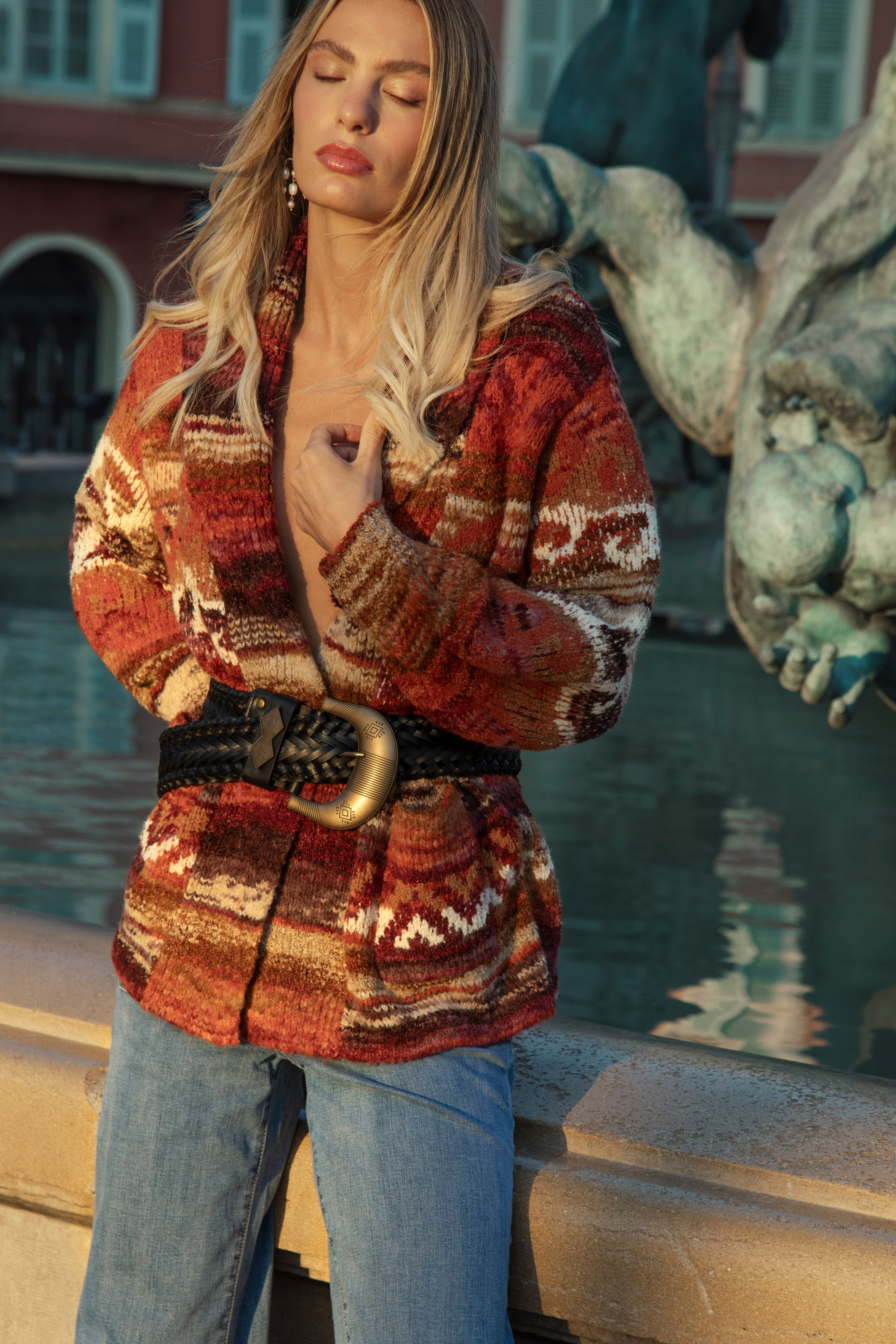 A woman wearing a rust-red and beige patterned knit cardigan with a black braided belt and gold buckle, paired with Paris Denim Jean