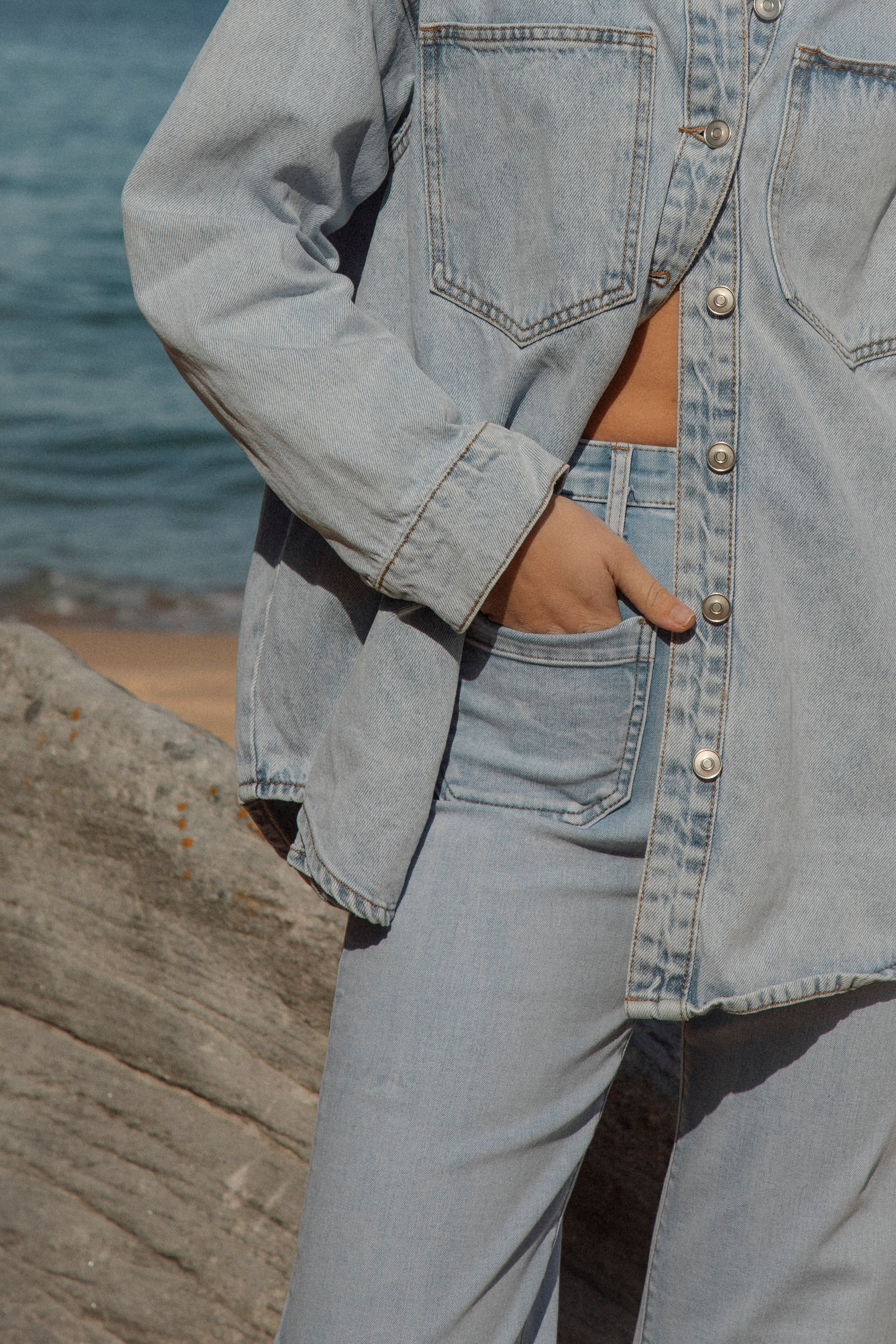 Woman wearing oversized light wash denim jacket with button front and chest pockets, styled with matching denim pants at the beach