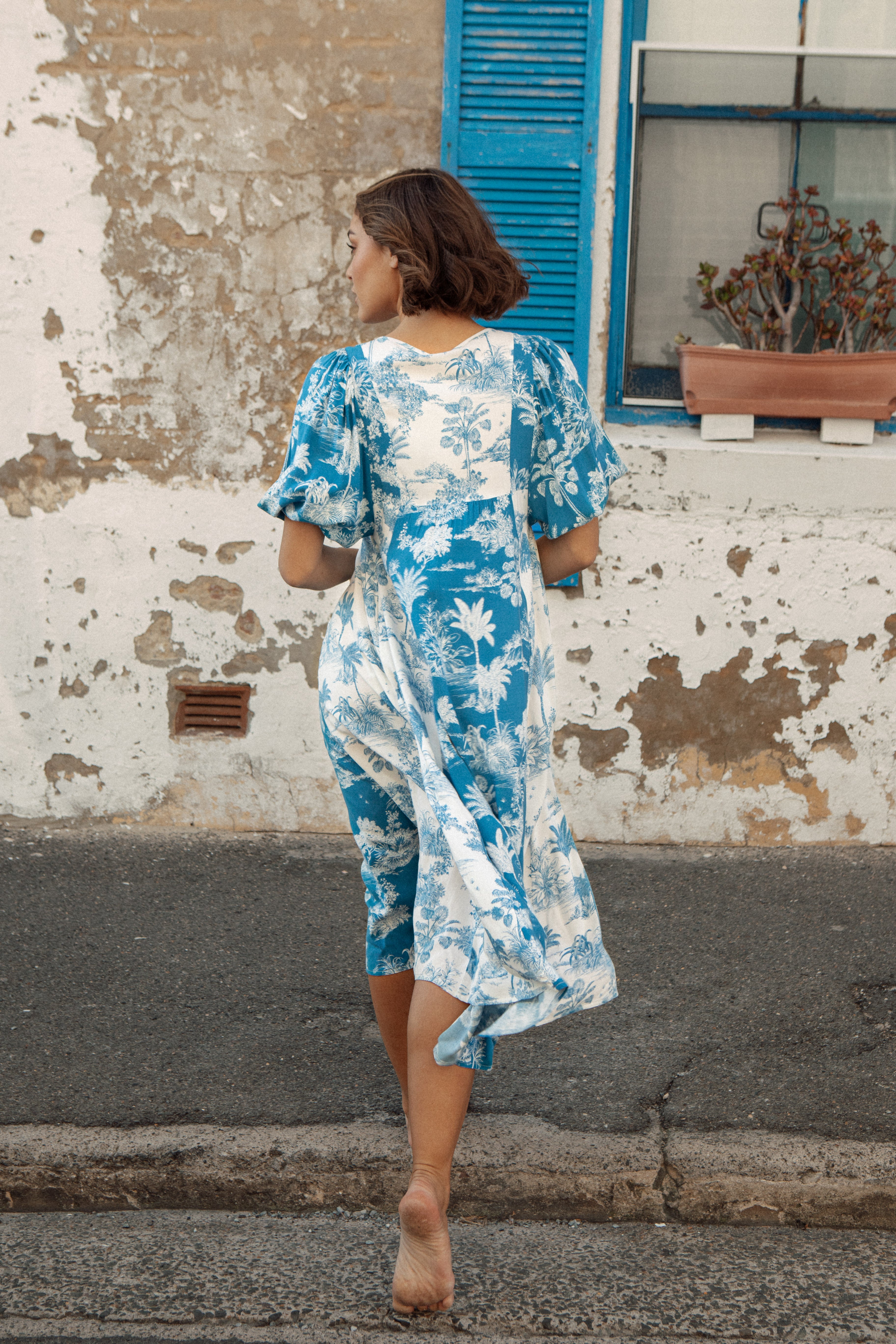 Woman in a blue and white floral dress walking on a street with a weathered wall and window in the background.